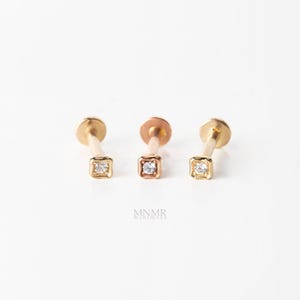 May include: Three gold stud earrings with flat circular backings and square-shaped heads, each featuring a single small clear gemstone set in the centre. The text MNMR is printed in small grey letters below the three earrings on a white background.