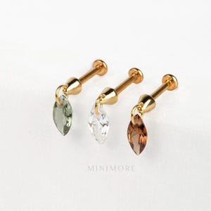 Może przedstawiać: Trzy złote kolczyki z wiszącymi, łezkowatymi kamieniami szlachetnymi w kolorze zielonym, przezroczystym i brązowym. Kolczyki mają sztyft i małe, okrągłe zapięcie. Na zdjęciu widnieje napis "MINIMORE".