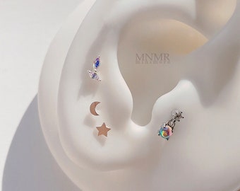 May include: A white ear model with five different earrings. The earrings are a crescent moon, a star, a small crystal, a larger crystal with a rainbow effect, and a small crystal with a rainbow effect.