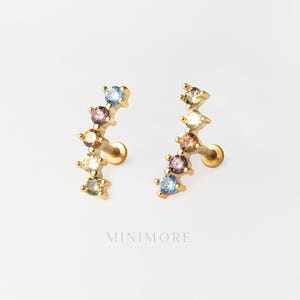 Puede incluir: Un par de pendientes dorados, cada uno con un diseño de barra curva engastado con cinco pequeñas piedras preciosas redondas multicolores en tonos azul, rosa, naranja y amarillo. La palabra MINIMORE está impresa debajo en letras mayúsculas.