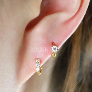 May include: Close-up of two gold hoop earrings with clear, sparkling gemstones. One earring features a single, larger stone, while the other has three smaller stones. The earrings are worn on an ear.