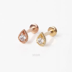 May include: Two stud earrings with flat back posts, one in rose gold and one in yellow gold. Each earring features a teardrop-shaped setting with a beaded border surrounding a single clear round gemstone. The text MNMR is visible below them.