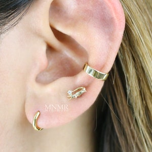 May include: Gold ear cuff with a small, clear gemstone. The ear cuff is worn on the upper part of the ear. A small gold hoop earring is worn in the lower lobe of the ear. The text "MNMR jewellery" is visible in the image.