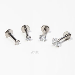 Puede incluir: Cuatro labrets plateados con gemas redondas y transparentes. Cada labret tiene una parte trasera plana en forma de disco y un poste con una gema engarzada con garras. Sobre un fondo blanco. Se ve el nombre de la marca "MNMR minimore".