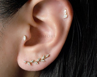 May include: A close-up of a person's ear with three gold earrings. The earrings are a small marquise-shaped diamond, a small leaf-shaped diamond, and a larger leaf-shaped earring with multiple diamonds.
