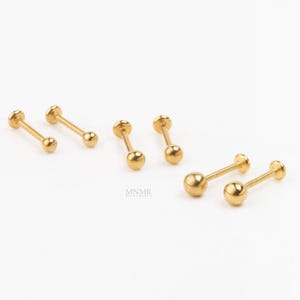 May include: A collection of gold-coloured barbell earrings, each with a ball at either end, are arranged against a white backdrop. The earrings are small and delicate, with a polished finish. The design is simple and minimalist.