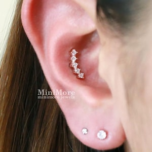 Puede incluir: Un pendiente de conch curvo en tono oro rosa con cinco pequeñas piedras transparentes de corte redondo dispuestas en arco vertical. La joya está situada en la zona del conch de la oreja, con dos pendientes de botón simples en el lóbulo.