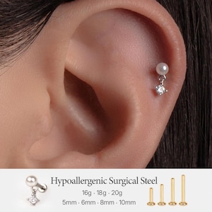 Peut inclure: Une boucle d'oreille en acier chirurgical hypoallergénique avec une perle blanche et une breloque en cristal transparent. Le texte indique Hypoallergenic Surgical Steel, 16g, 18g, 20g, 5mm, 6mm, 8mm, 10mm. Le bijou est porté sur le cartilage de l'oreille.