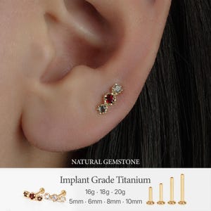May include: A gold-tone stud earring featuring three prong-set stones in a row, with a central deep red stone flanked by two clear stones. Text includes NATURAL GEMSTONE, Implant Grade Titanium, 16g, 18g, 20g, 5mm, 6mm, 8mm, and 10mm.