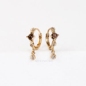 May include: A pair of gold-coloured hoop earrings. Each earring has a brown, square-shaped stone, smaller clear stones, and a dangling clear stone. The word "MINIMORE" is printed on the bottom.