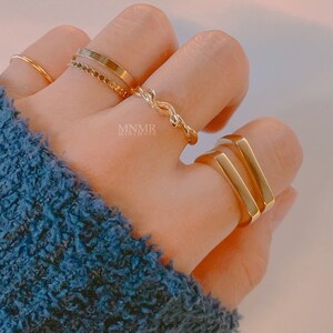 May include: A set of gold rings, including a thin band, a chain ring, a twisted ring, and a double bar ring. The rings are worn on a hand.