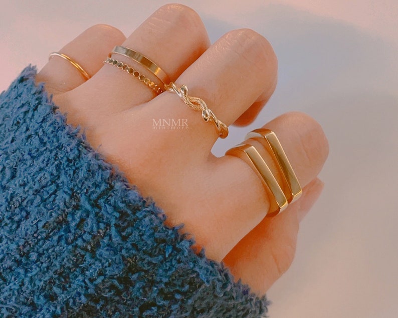 May include: A set of gold rings, including a thin band, a chain ring, a twisted ring, and a double bar ring. The rings are worn on a hand.