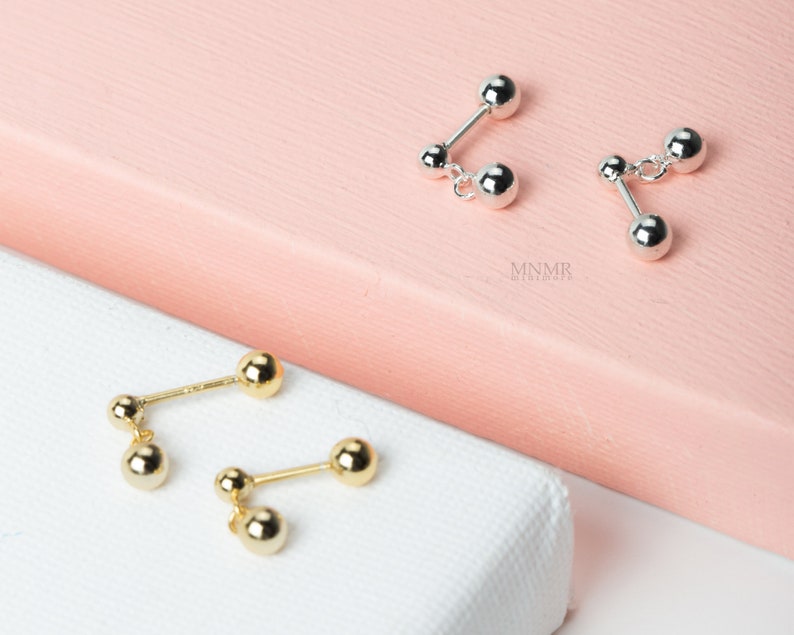 May include: Two pairs of earrings, one pair is gold and the other is silver. Both pairs have two small balls connected by a bar.