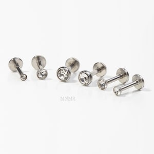 May include: A collection of six silver-colored, flat-backed labret studs. Each stud features a small, clear, round gemstone. The studs vary in size, with some having a longer post and smaller gem, and others a shorter post and larger gem. The brand name "MNMR" is at the bottom.