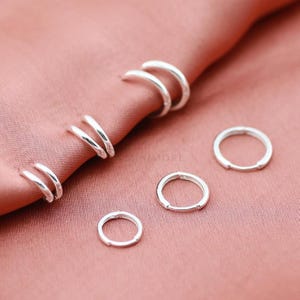 May include: A collection of small, polished silver hoop earrings. Three pairs are partially tucked into the folds of a dusty rose fabric, while three individual silver hoop earrings are arranged separately on the surface below the fabric.