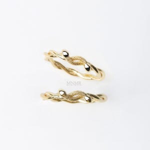 Può includere: Due anelli color oro con un design intrecciato e piccoli accenti arrotondati. Gli anelli sono esposti su uno sfondo bianco, mettendo in risalto i loro dettagli intricati e la finitura lucida. Gli anelli fanno probabilmente parte di una collezione di gioielli.