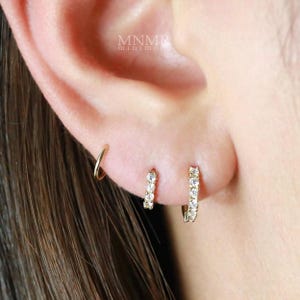 May include: Close-up of three gold earrings on an ear. The first is a small hoop, the second is a small hoop with three clear stones, and the third is a larger hoop with multiple clear stones. The brand name "MNMR minimore" is visible.