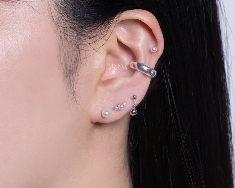 May include: A person's ear with multiple silver earrings. The earrings include a hoop, a small pearl stud, and a dangling pearl earring.