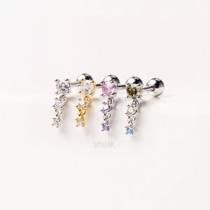 May include: Four silver helix earrings with dangling gemstones in clear, gold, pink, and green. Each earring has a post and ball closure. The earrings are adorned with a cascade of small, colourful gems. The brand name "MNMR" is printed in the center.