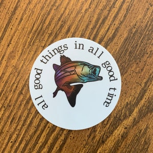 May include: A circular sticker featuring a colourful fish illustration. The fish is depicted in shades of orange, blue, and purple. The text "all good things in all good time" is printed around the fish in a circular pattern.