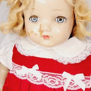 Puede incluir: Una muñeca de porcelana vintage con cabello rubio que lleva un vestido rojo con ribete de encaje blanco y un cuello blanco.