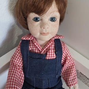 Puede incluir: Una muñeca de porcelana con cabello corto castaño rojizo, ojos azules y pecas. La muñeca viste una camisa de cuello a cuadros rojos y blancos debajo de un peto de mezclilla azul oscuro. Tiene una tez pálida y una expresión facial neutra.