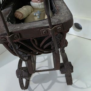 Puede incluir: Una muñeca antigua en un cochecito de bebé de madera vintage. El cochecito es de color marrón oscuro con detalles metálicos y ruedas de radios. La muñeca lleva un suéter color crema con un adorno azul.