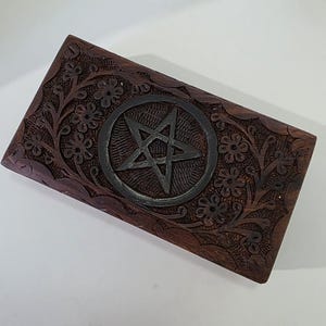May include: A rectangular, dark brown wooden box with intricate floral carvings and a central pentagram design. The pentagram is set within a circular frame, adding a contrasting element to the box's overall aesthetic.