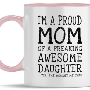 May include: White ceramic coffee mug with a pink rim and handle. The mug features black text that reads "I'm a proud mom of a freaking awesome daughter...yes, she bought me this."