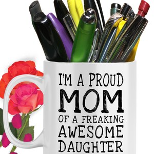 May include: White ceramic coffee mug with a black handle. The mug has black text that reads "I'm a proud mom of a freaking awesome daughter...yes, she bought me this".