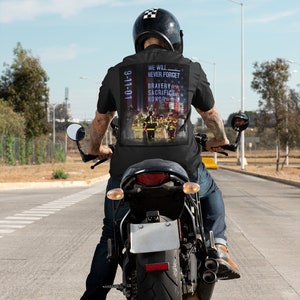 Pu&ograve; includere: Un uomo indossa una maglietta nera con una grafica 9-11-01 sul retro. La grafica presenta una rappresentazione di vigili del fuoco con le parole "We Will Never Forget" e "Bravery Sacrifice Honor". L'uomo sta guidando una motocicletta nera su una strada asfaltata.