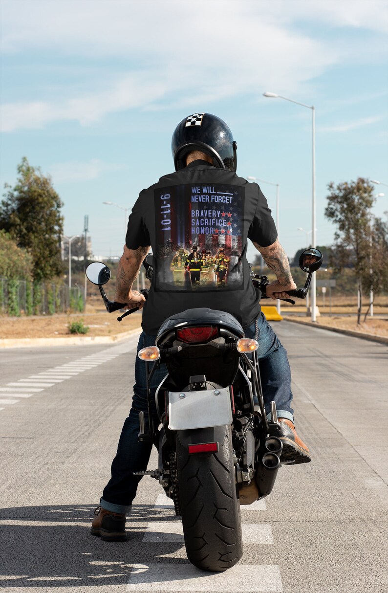 Pu&ograve; includere: Un uomo indossa una maglietta nera con una grafica 9-11-01 sul retro. La grafica presenta una rappresentazione di vigili del fuoco con le parole "We Will Never Forget" e "Bravery Sacrifice Honor". L'uomo sta guidando una motocicletta nera su una strada asfaltata.