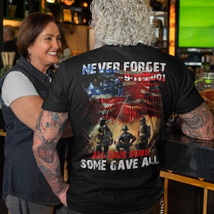 Pu&ograve; includere: Una maglietta nera con una grafica di vigili del fuoco di fronte alle Torri Gemelle con una bandiera americana drappeggiata sulle torri. Il testo "Never Forget 9-11-2001" &egrave; sopra la grafica e il testo "All Gave Some Some Gave All" &egrave; sotto la grafica.