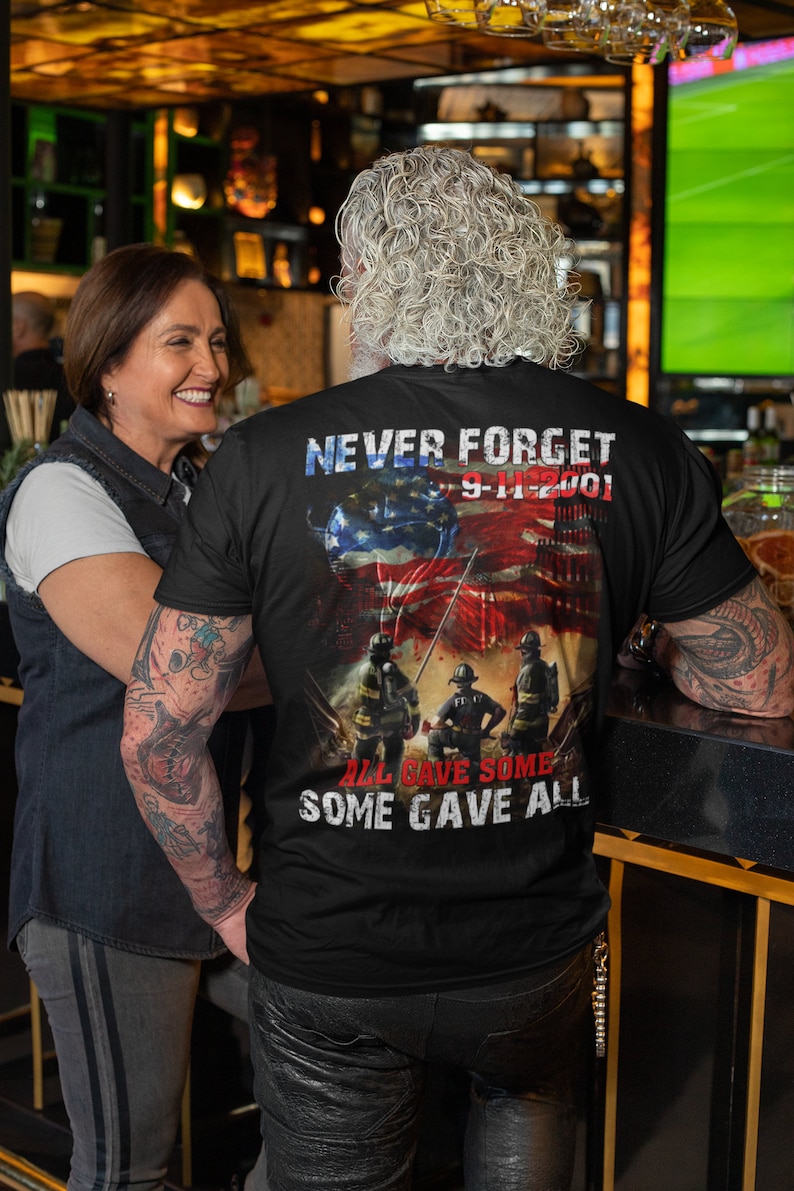 Pu&ograve; includere: Una maglietta nera con una grafica di vigili del fuoco di fronte alle Torri Gemelle con una bandiera americana drappeggiata sulle torri. Il testo "Never Forget 9-11-2001" &egrave; sopra la grafica e il testo "All Gave Some Some Gave All" &egrave; sotto la grafica.