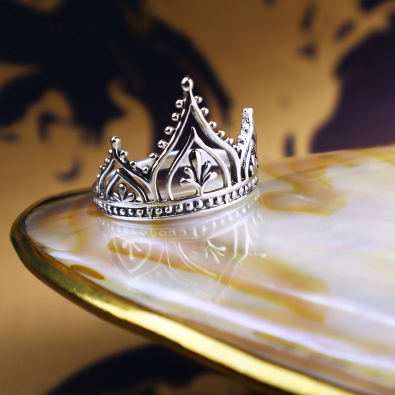 K&ouml;nnte beinhalten: Ein silberner Kronenring mit einem filigranen, komplizierten Design. Der Ring liegt auf einer wei&szlig;en und goldenen muschelf&ouml;rmigen Oberfl&auml;che.