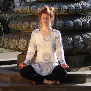 Puede incluir: Una mujer se sienta con las piernas cruzadas en meditación, vistiendo una túnica blanca con un patrón azul y blanco. La túnica tiene cuello en V y mangas largas. La mujer tiene el pelo rojo y lleva una camisa blanca debajo de la túnica. Está sentada frente a una fuente grande y ornamentada.
