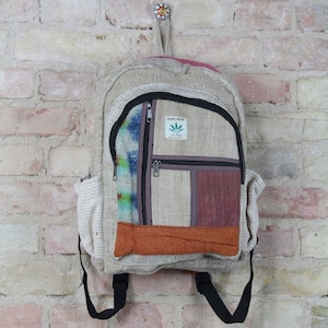 May include: A beige hemp backpack with a brown, orange, and purple patchwork design. The backpack has a zippered pocket on the front with a label that reads "PURE HEMP THE FINE FIBER MADE IN NEPAL".