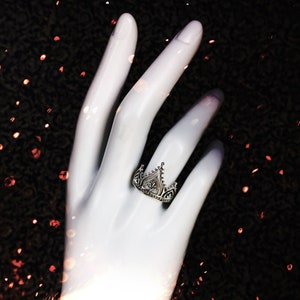 K&ouml;nnte beinhalten: Ein silberner Ring mit einem Kronendesign. Der Ring befindet sich an einem wei&szlig;en Handmodell vor einem schwarzen Hintergrund mit rotem Glitzer.