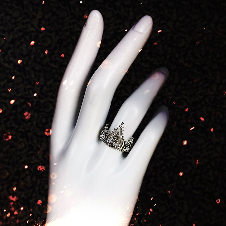 K&ouml;nnte beinhalten: Ein silberner Ring mit einem Kronendesign. Der Ring befindet sich an einem wei&szlig;en Handmodell vor einem schwarzen Hintergrund mit rotem Glitzer.