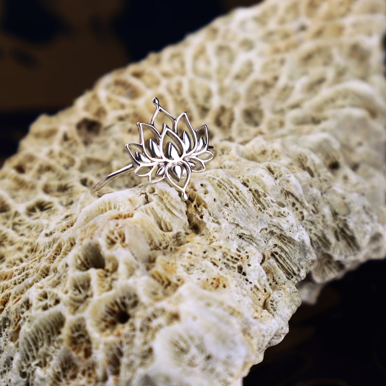 Puede incluir: Un anillo de plata con un delicado dise&ntilde;o de flor de loto. El anillo se muestra sobre una superficie blanca texturizada.