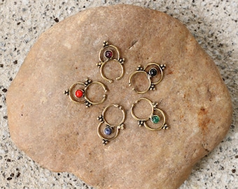 Puede incluir: Seis anillos de nariz de oro con piedras preciosas de diferentes colores. Los anillos están dispuestos en círculo sobre una roca marrón.