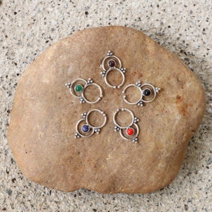 Könnte beinhalten: Fünf silberne Nasenringe mit verschiedenen Edelsteinen. Die Ringe sind in einem Kreis auf einem braunen Stein angeordnet.