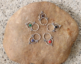 Puede incluir: Cinco anillos de nariz de plata con piedras preciosas de diferentes colores. Los anillos están dispuestos en círculo sobre una roca marrón.