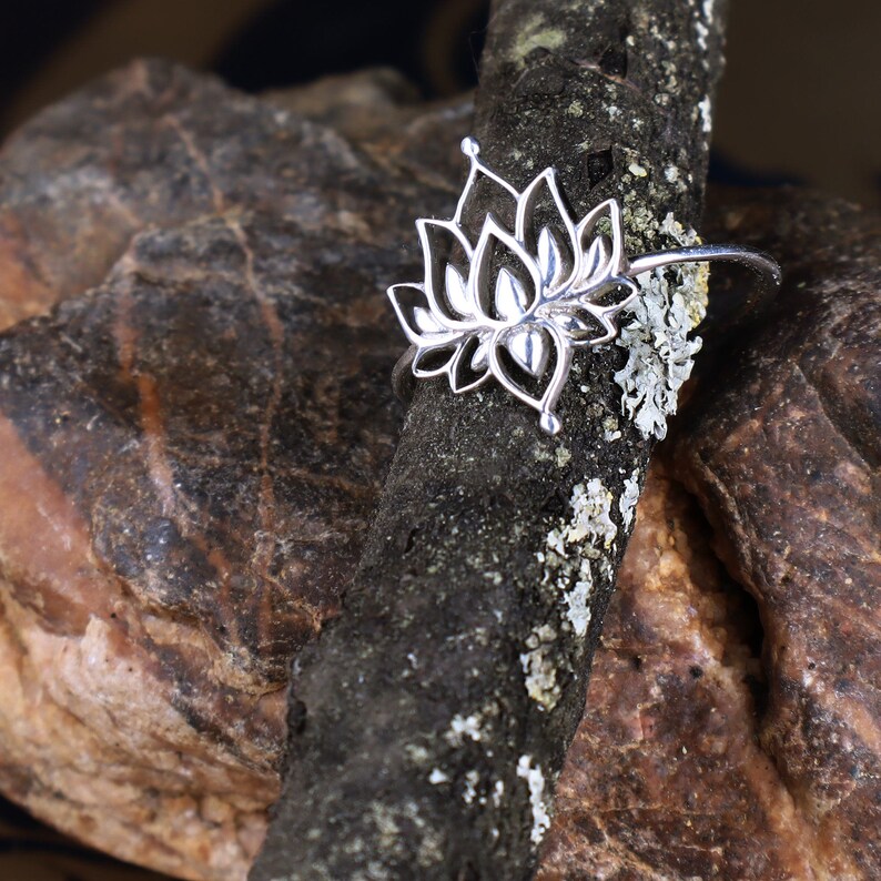 Puede incluir: Un anillo de plata con un delicado dise&ntilde;o de flor de loto. El anillo se muestra en un trozo de madera con una textura rugosa.