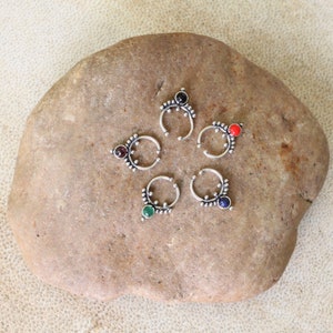 Könnte beinhalten: Fünf silberne Nasenringe mit verschiedenen Edelsteinen. Die Ringe sind in einem Kreis auf einem grauen Stein angeordnet.