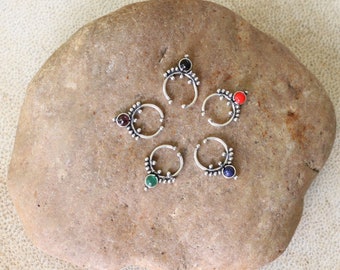Puede incluir: Cinco anillos de nariz de plata con piedras preciosas de diferentes colores. Los anillos están dispuestos en círculo sobre una roca gris.