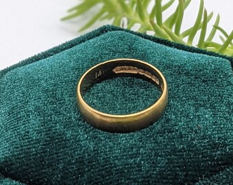 May include: A gold ring in a dark green velvet ring box. The box is hexagonal and has a textured surface. The ring is simple and has a polished finish.