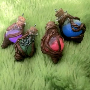 Puede incluir: Cuatro botellas de poción en miniatura con tapones de corcho. Cada botella contiene un líquido de diferente color: morado, verde, rojo y azul. Las botellas están envueltas en arcilla marrón, lo que les da un aspecto rústico y hecho a mano. El fondo es una superficie verde borrosa.
