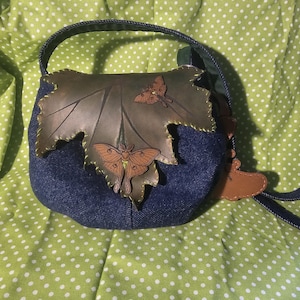 May include: A denim handbag with a leaf-shaped leather applique. The leaf is brown with stitched edges and features three brown moth appliques. The bag has a blue denim body, a dark blue handle, and a green lining.