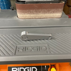 May include: A gray and orange Ridgid belt sander with a gray lever handle. The sander has a brown sanding belt and a black and orange dust collection port. The sander is on a gray surface with a black and white striped pattern.